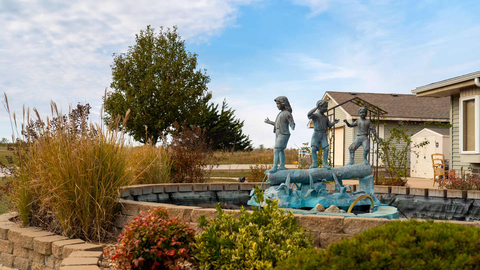 Outdoor garden area with a decorative water feature showing a statue of three children playing on a log surrounded by plants and shrubs, with a building and a tree in the background under a partly cloudy sky.