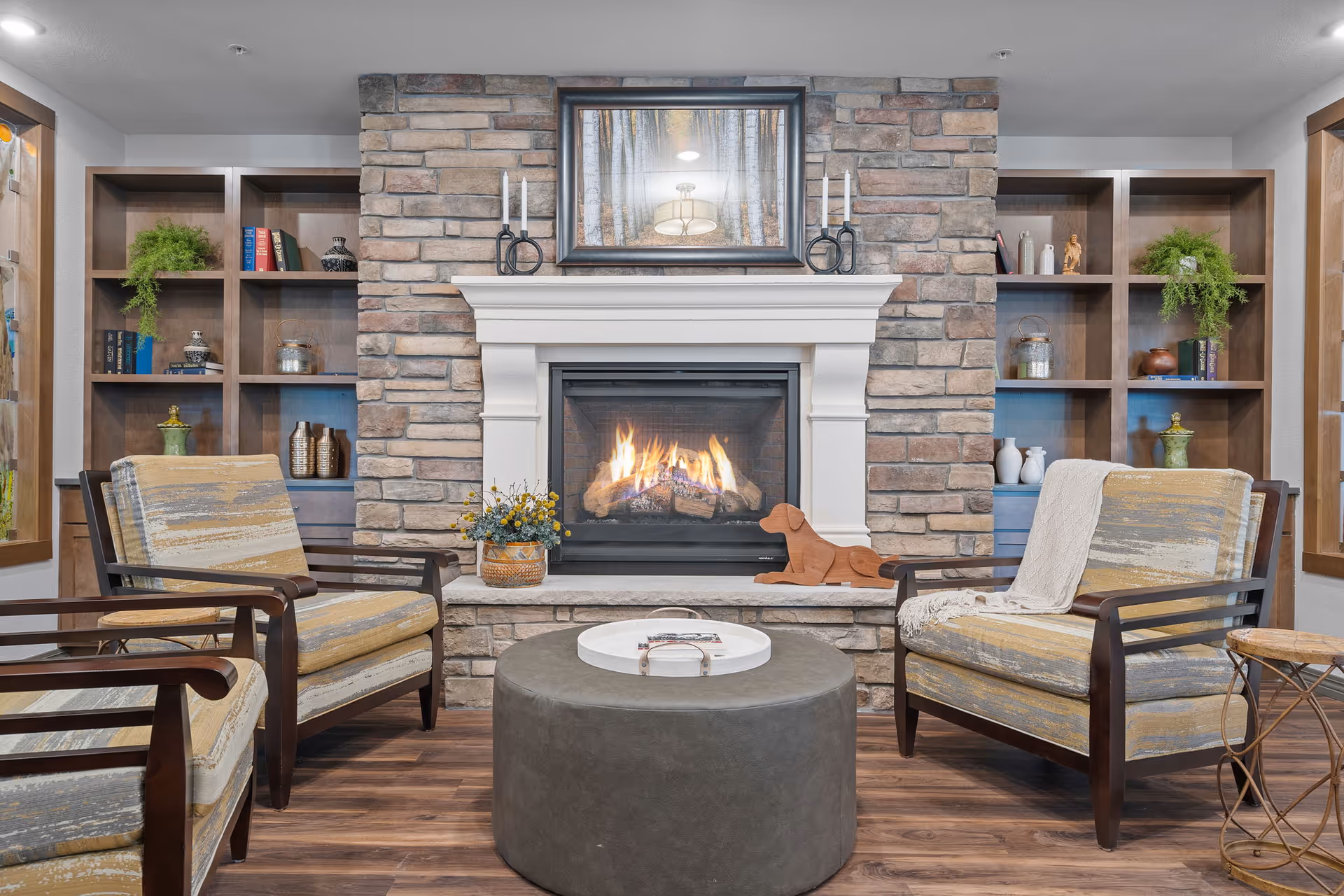 Cozy communal living room with a stone fireplace flanked by built-in shelves, two armchairs and a round ottoman.