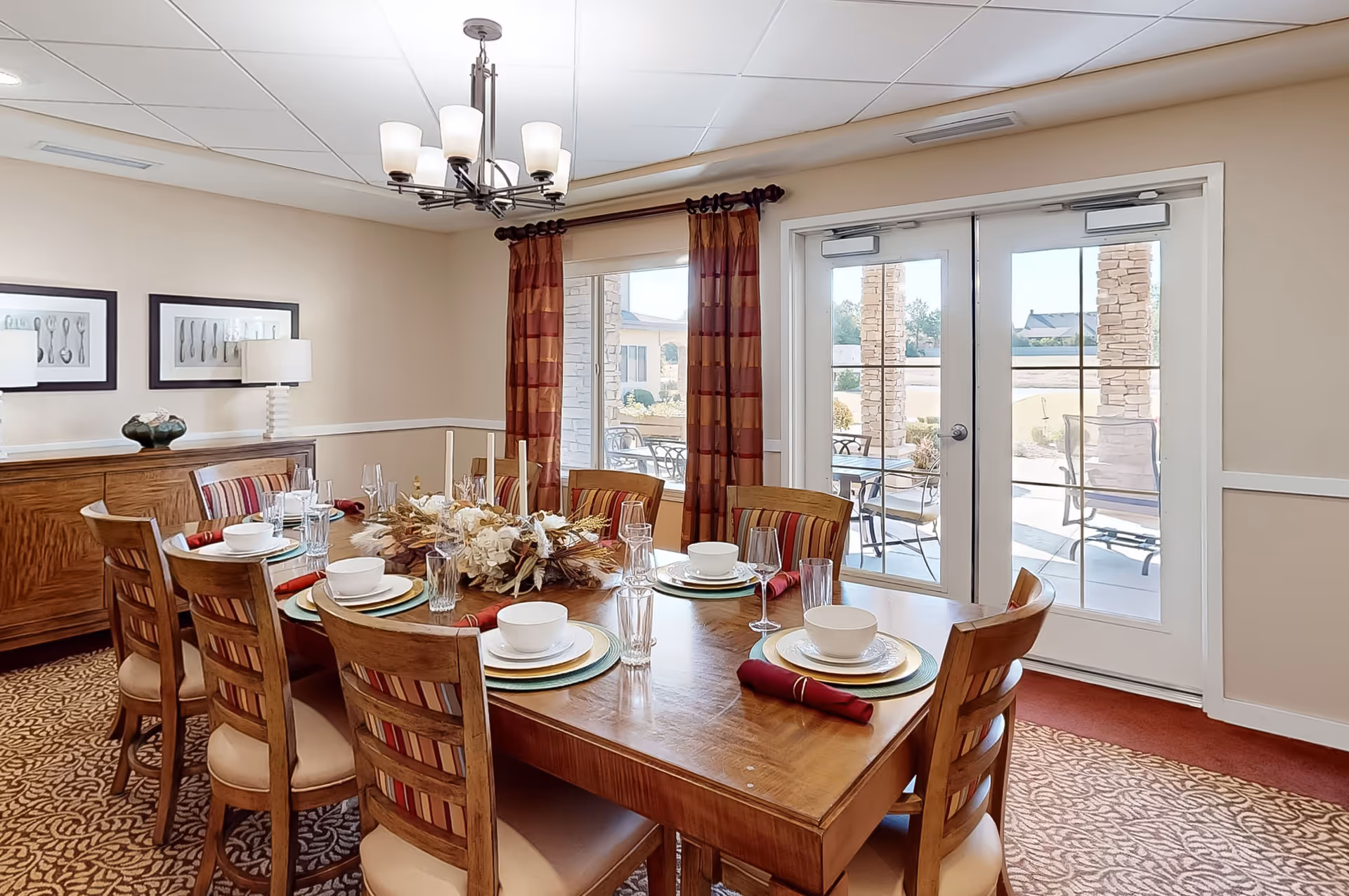 A dining room with a wooden table set for eight people, featuring white bowls, plates, glasses, and red napkins. The chairs have wooden frames with striped upholstery. A floral centerpiece decorates the table. The room has beige walls, a patterned carpet, two framed pictures on the wall, a sideboard with lamps, and large windows with red and orange striped curtains. Double glass doors lead to an outdoor patio area with tables and chairs.