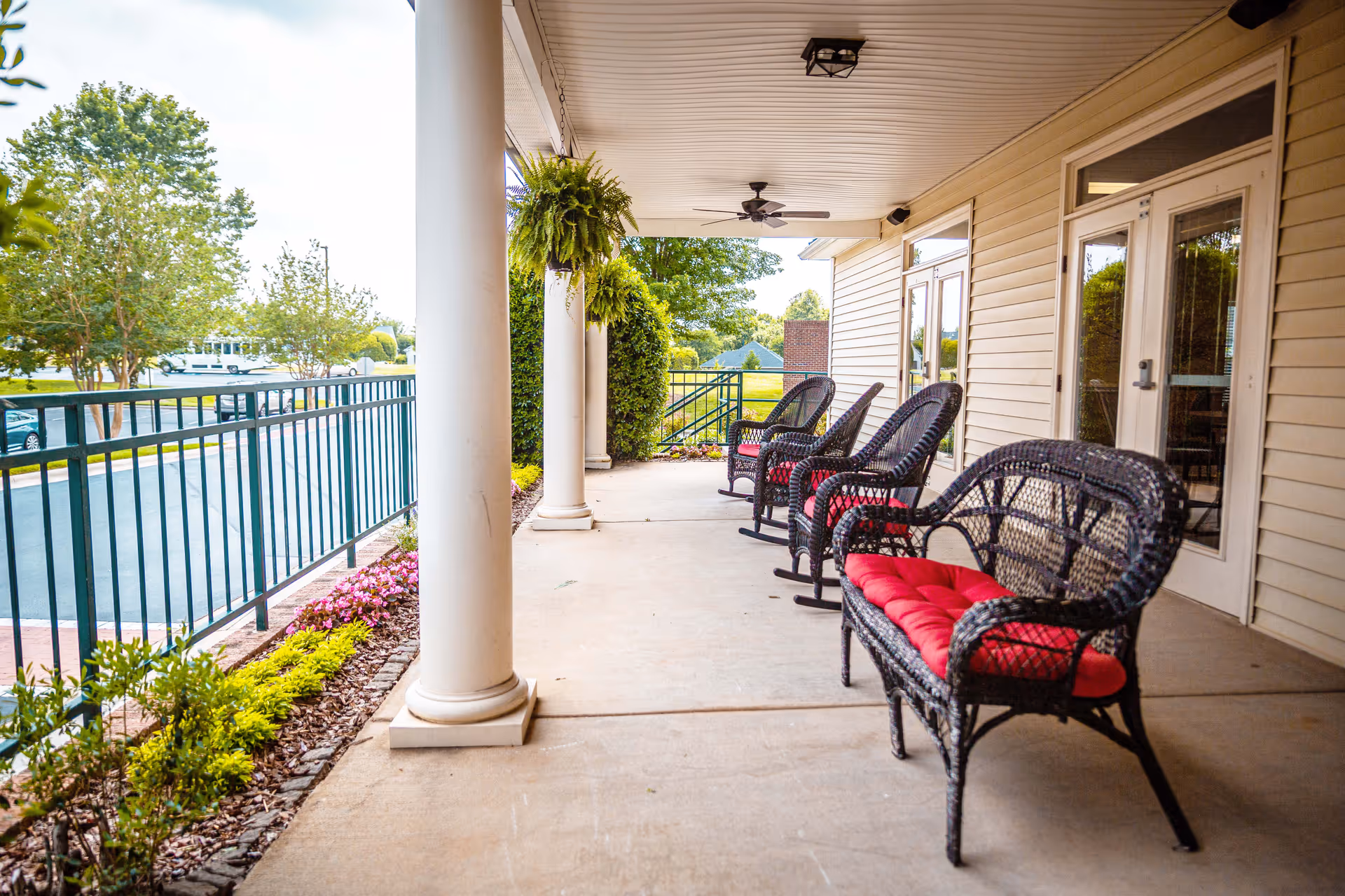Covered outdoor patio area with black wicker rocking chairs and a wicker bench, all with red cushions. The patio has white columns, hanging green plants, ceiling fans, and overlooks a parking lot and landscaped garden area.