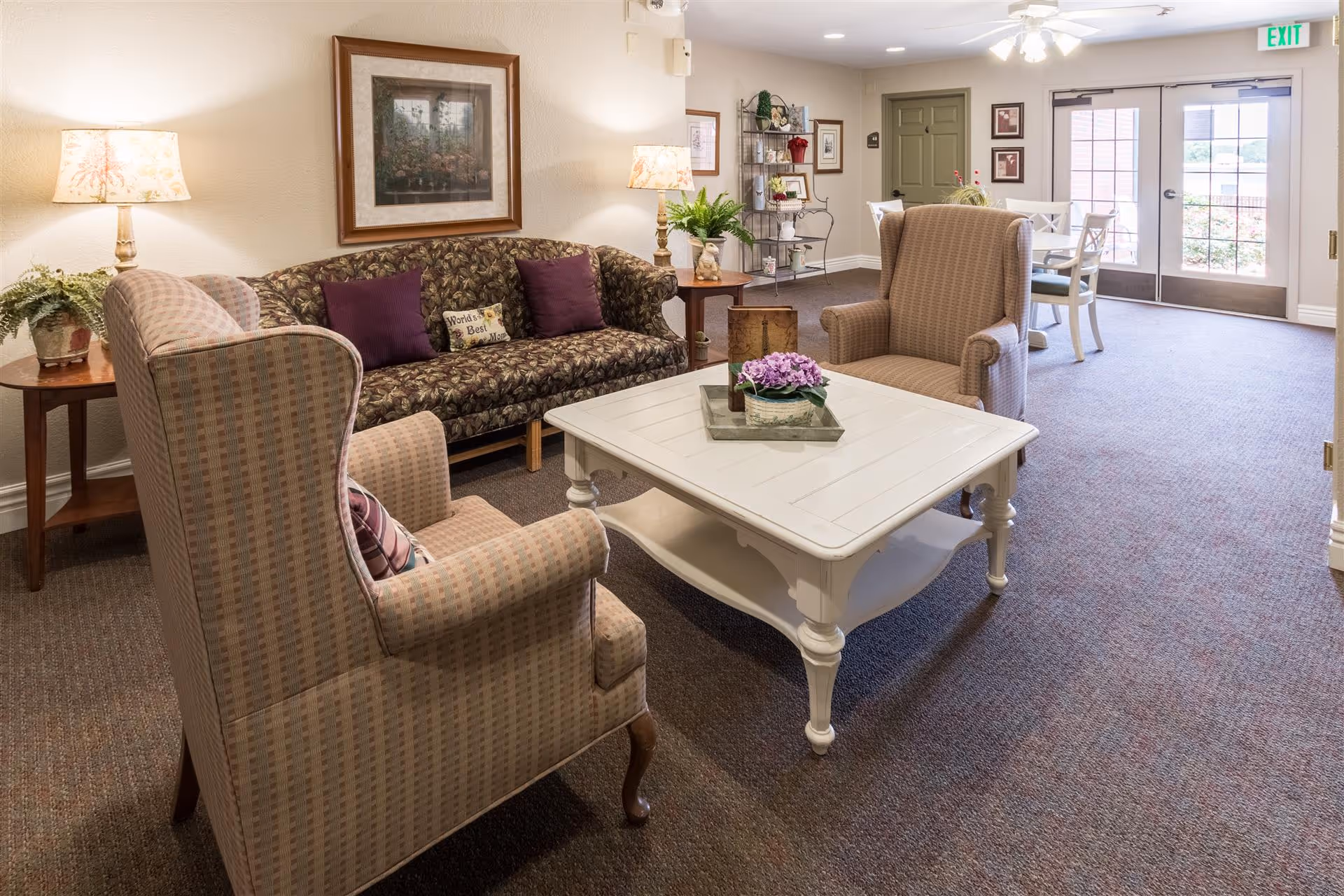 Seating area in a senior living facility with armchairs, a patterned sofa, coffee table, lamps, and a dining area by glass doors.
