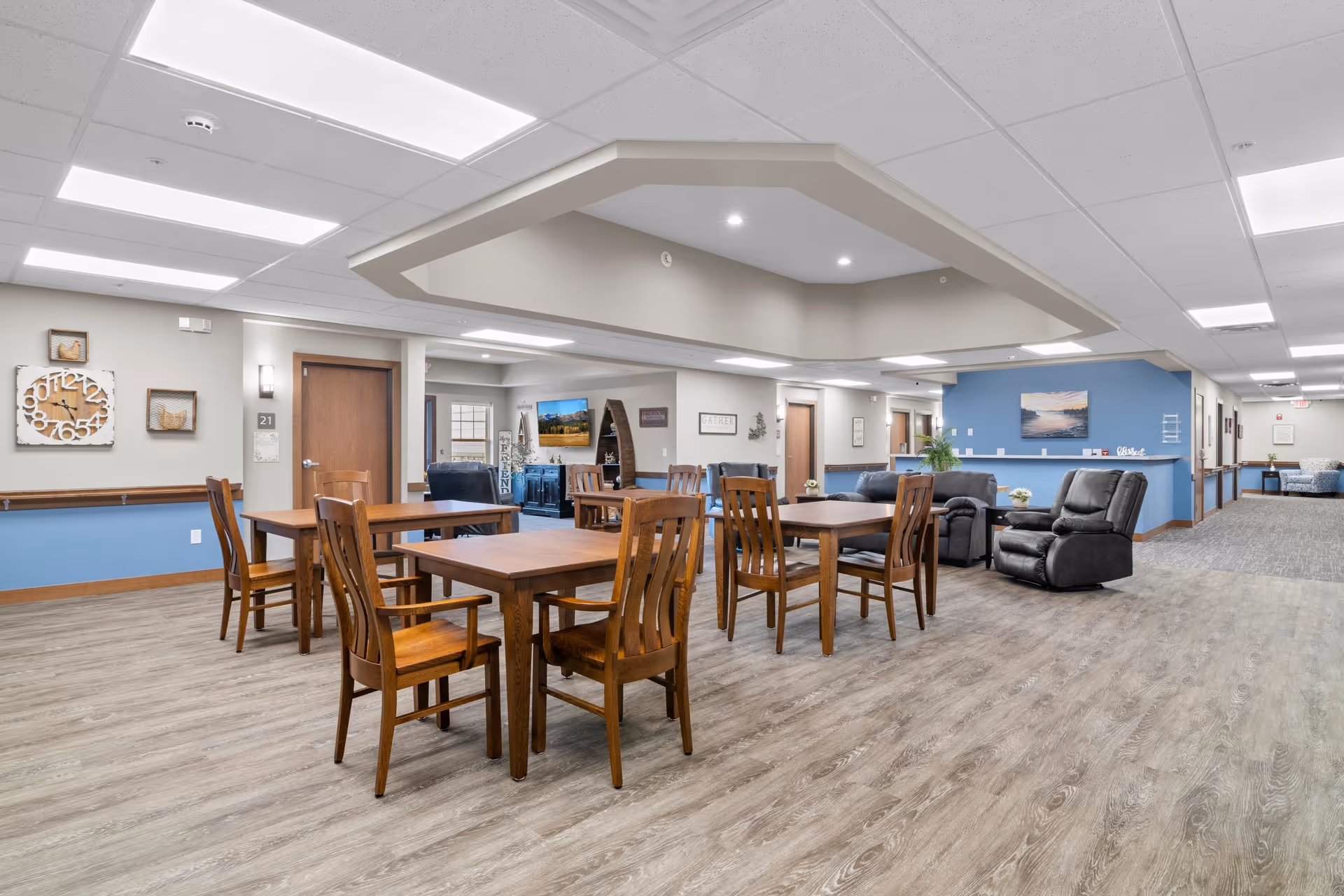 A spacious and well-lit common area in a senior living facility featuring wooden tables and chairs arranged for dining or socializing. The room has light-colored walls with blue accents, a decorative ceiling feature with recessed lighting, comfortable recliner chairs, and artwork on the walls. The flooring is wood-style laminate, and there are multiple doorways leading to other rooms or hallways.