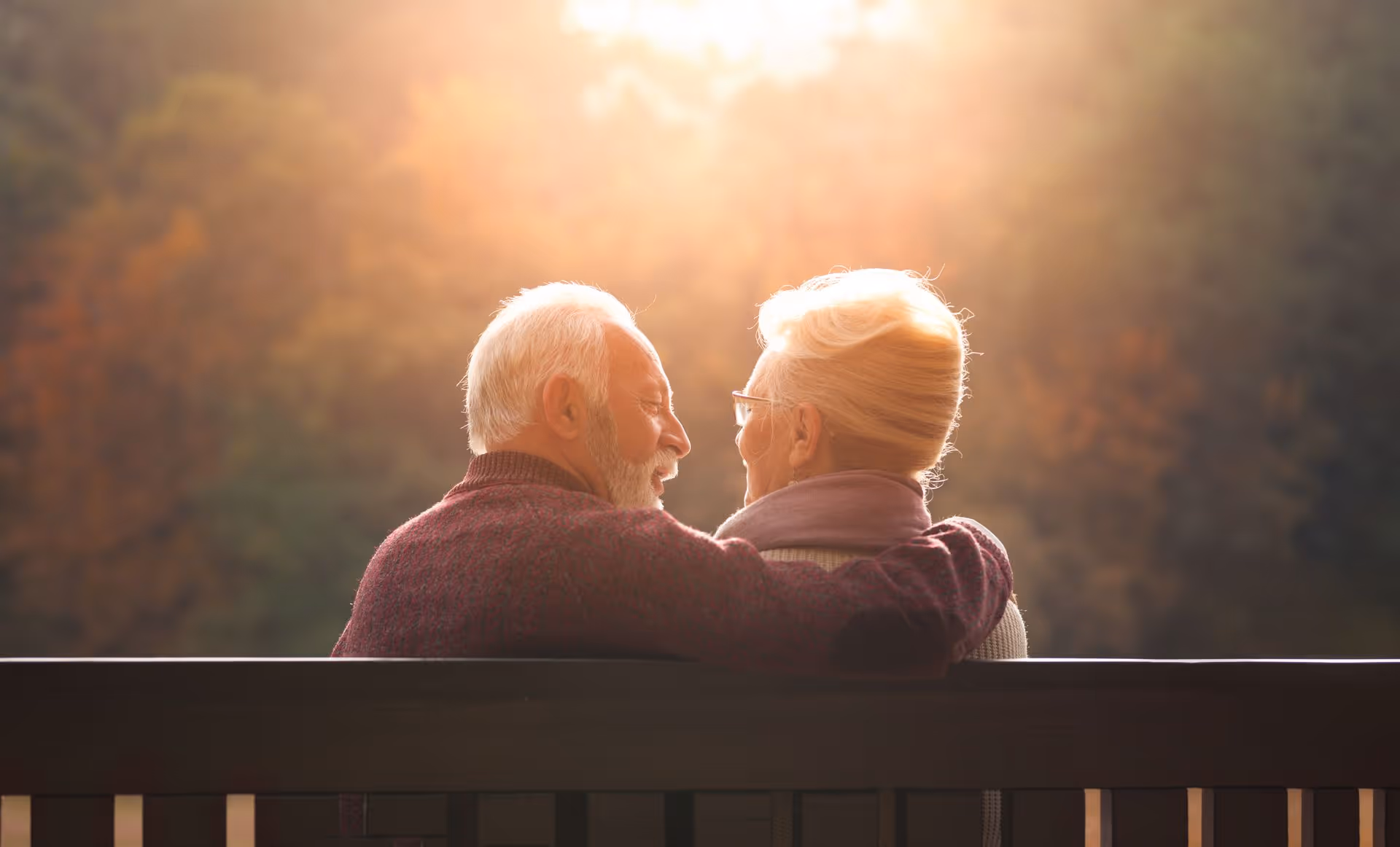 An elderly couple sitting closely together on a bench outdoors, with the man’s arm around the woman, bathed in warm sunlight and surrounded by blurred autumn foliage.