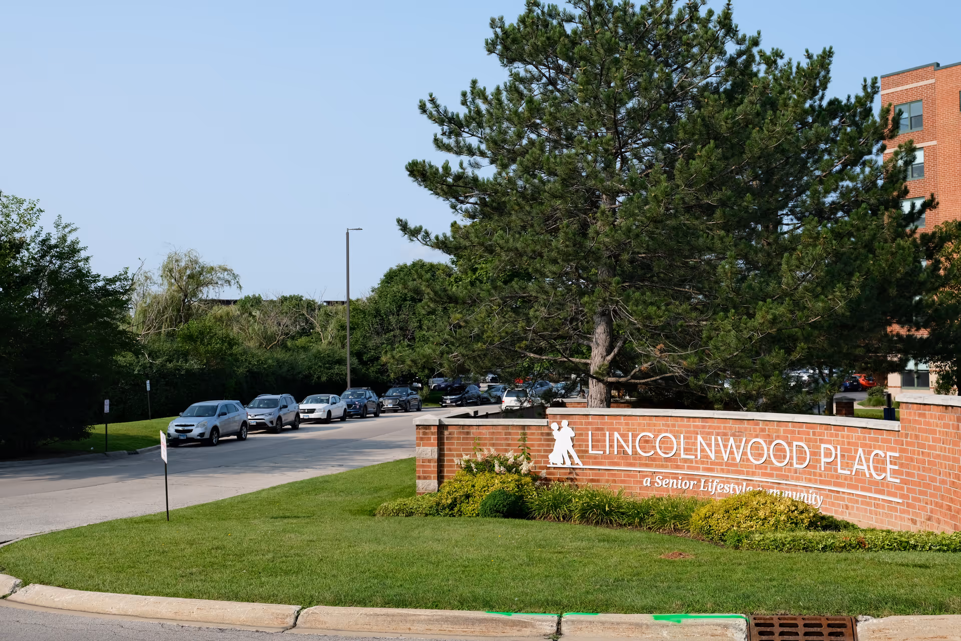 Entrance sign for Lincolnwood Place, a senior lifestyle community, displayed on a brick wall with landscaping and a large tree nearby. Several cars are parked along the road next to the sign, and part of a multi-story brick building is visible in the background under a clear blue sky.