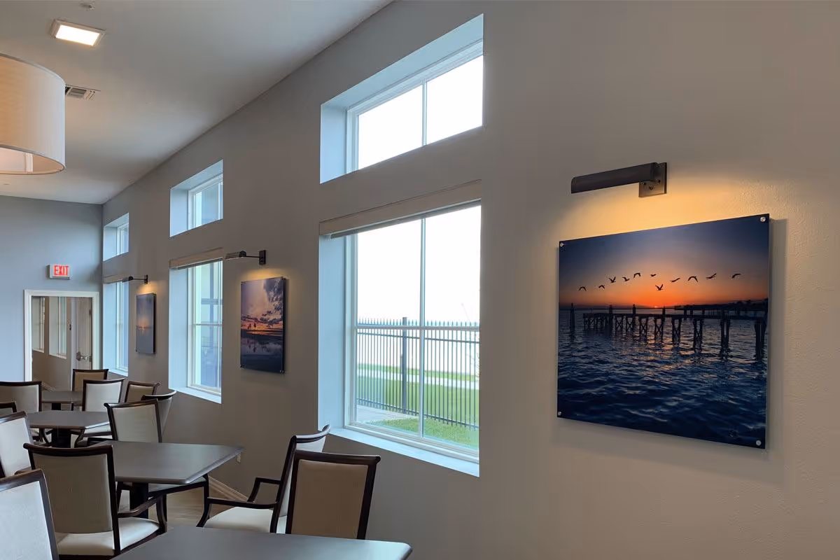 Interior view of a dining area with multiple tables and chairs arranged neatly. The room has large windows letting in natural light and framed pictures of sunset scenes with birds on the walls, each illuminated by individual wall lights. A lamp hangs from the ceiling and an exit sign is visible in the background.