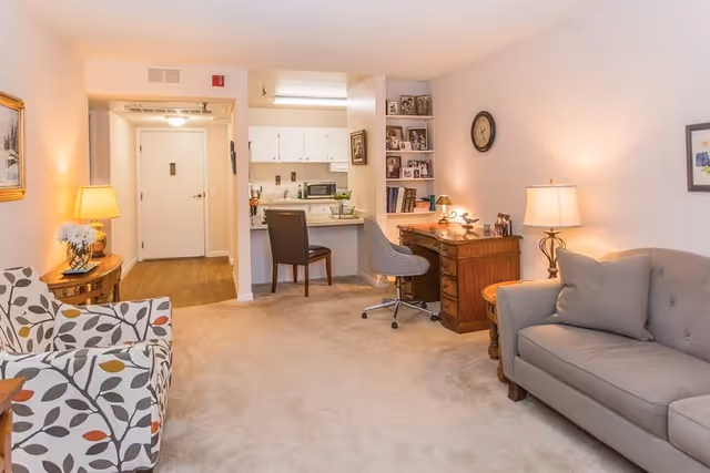 Cozy living room with a sofa and patterned armchair, a wooden desk workspace, and a small open kitchenette in the background.