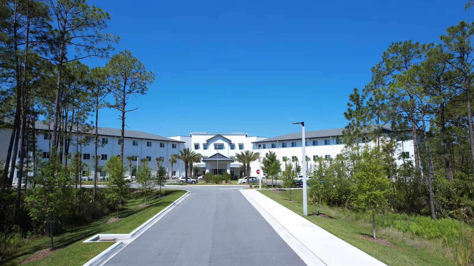 Front exterior view of a large, white, three-story assisted living and memory care facility named Paradise Pointe Assisted Living & Memory Care, surrounded by trees and greenery under a clear blue sky.