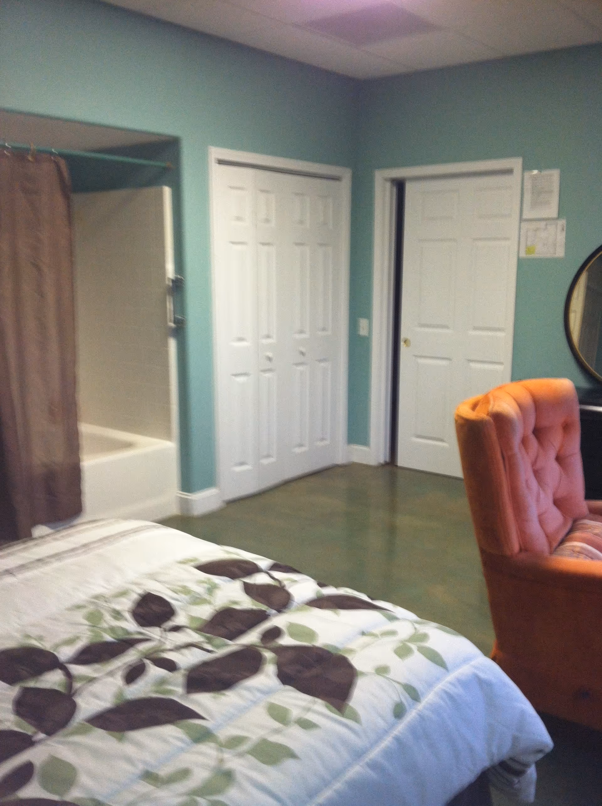 Bed with a patterned duvet in the foreground, an orange upholstered chair, white closet doors, and a bathtub/shower alcove along teal-painted walls.