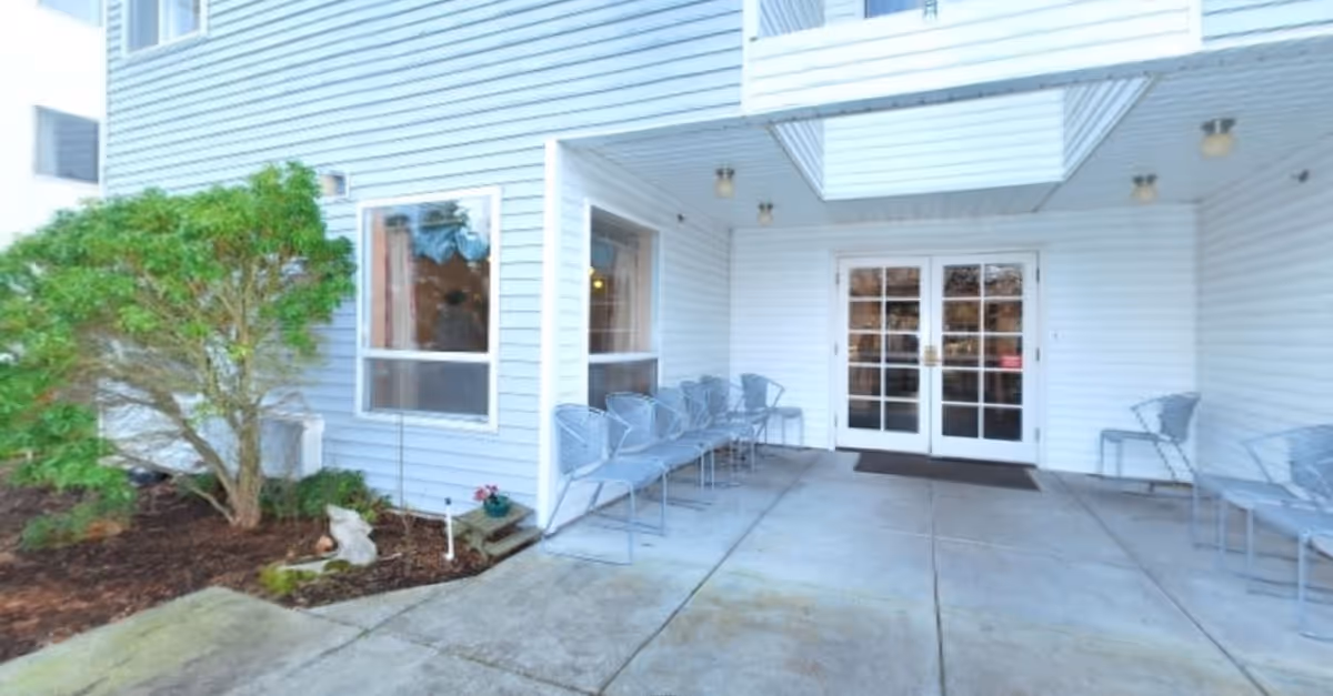 Covered outdoor seating area with metal chairs arranged along the walls near a white building entrance with double glass doors. There is a small garden with a bush and some plants to the left of the seating area.
