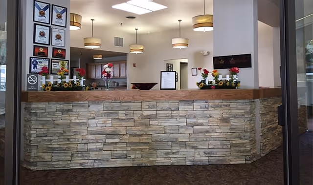 Reception desk area inside Madison House-Assisted Living with a stone facade and wooden countertop. The desk is decorated with small flower arrangements and candle holders. Behind the desk, there are several hanging ceiling lights, framed awards on the wall to the left, and a person partially visible sitting behind the desk.