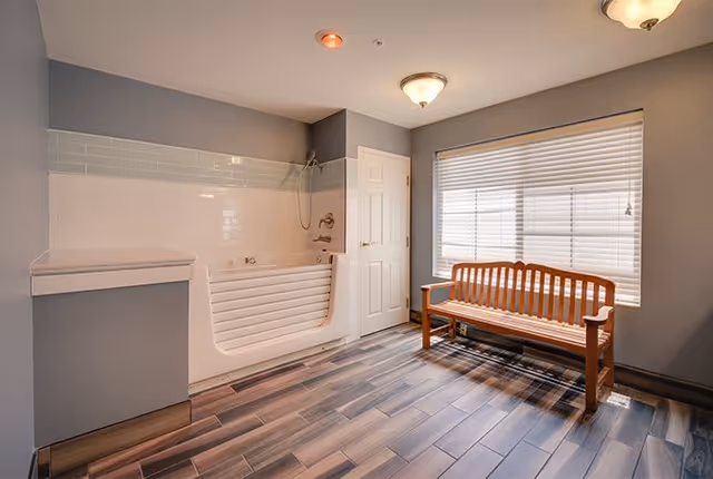 A spacious bathroom with a walk-in bathtub on the left side, a wooden bench near a large window with blinds, and wood-patterned tile flooring. The walls are painted gray with white tiles around the bathtub area. There is a ceiling light fixture and a closed white door next to the bathtub.