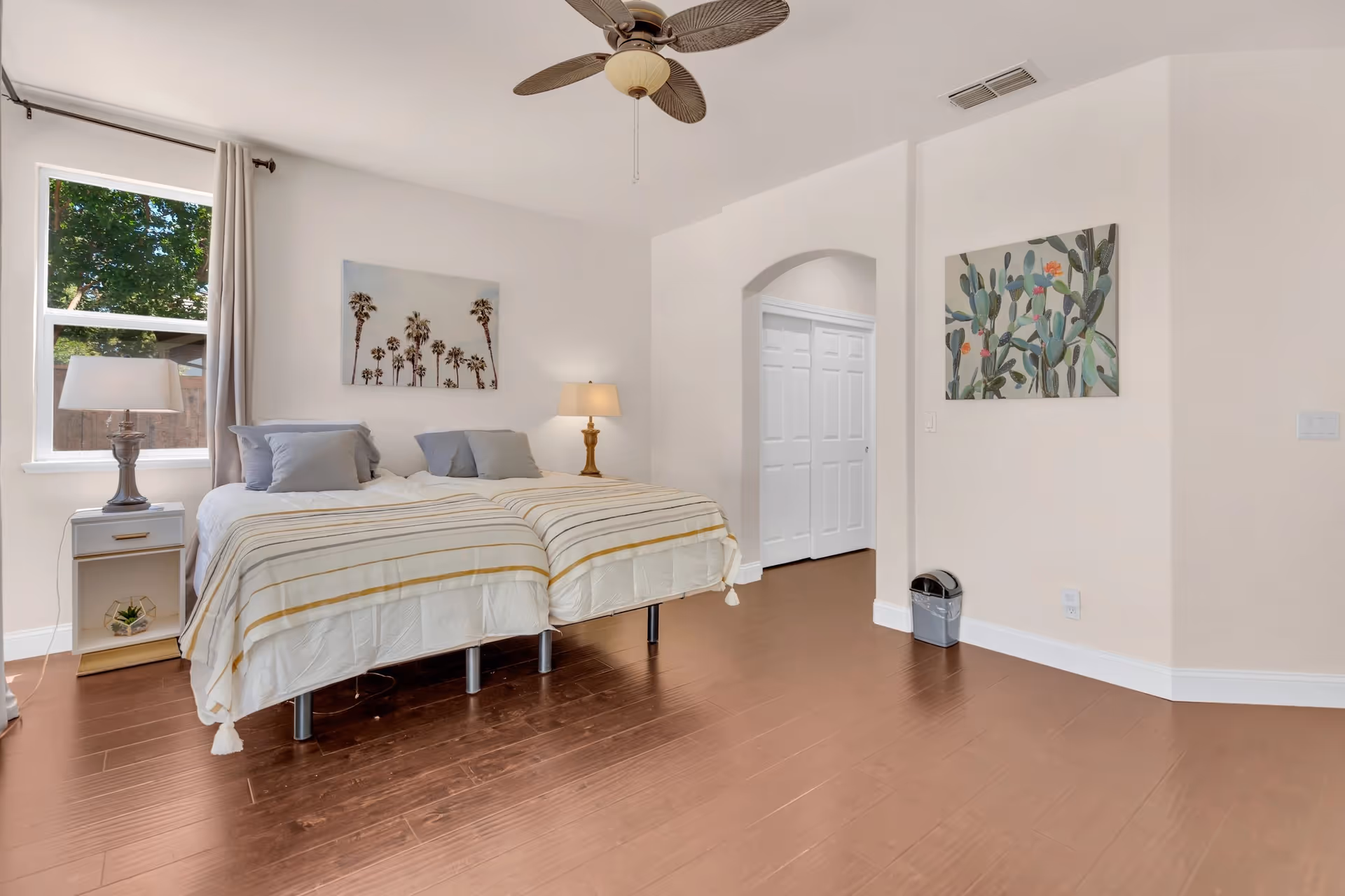Bright bedroom with two twin beds pushed together, nightstands, lamps, ceiling fan, hardwood floors, and wall art.