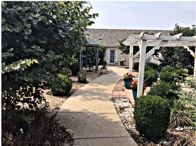 A paved courtyard walkway leading past pergolas, potted flowers, and trimmed shrubs toward a building entrance.