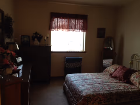A bedroom with a floral patterned bedspread on a bed, two matching pillows, a wooden dresser with a mirror, a tall chest of drawers, a window with a red valance curtain, and a standing mirror in the corner.