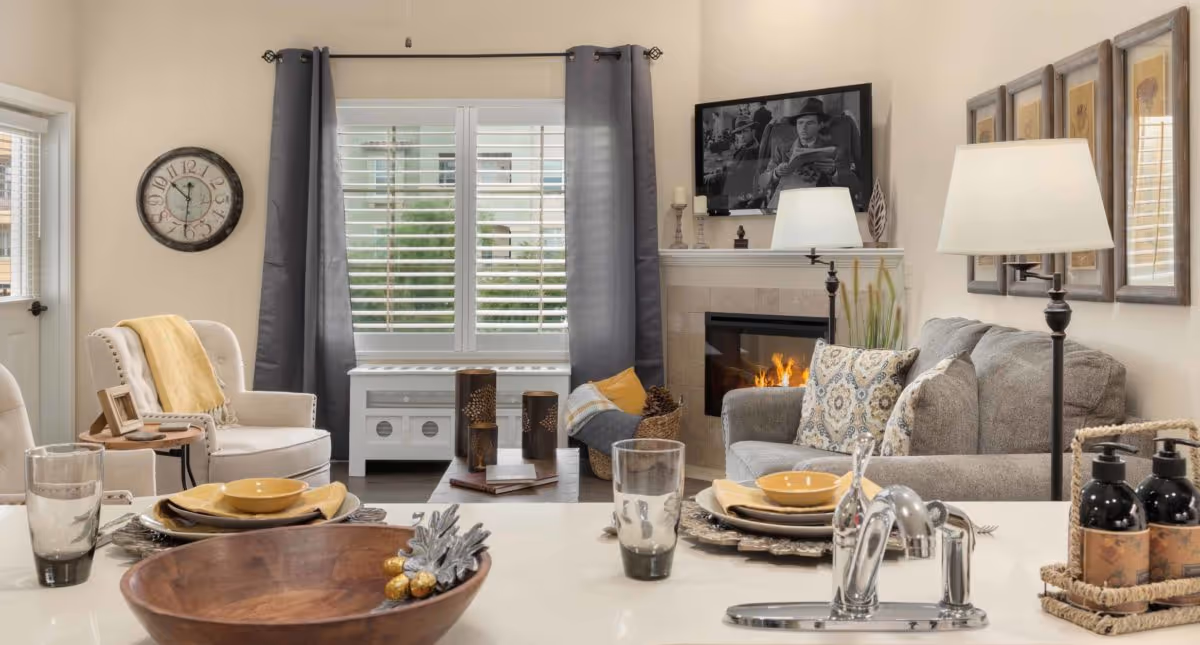 A cozy living room with a gray sofa adorned with patterned cushions, a lit fireplace, and a black-and-white framed photo above the mantle. There is a white armchair with a yellow throw blanket, a round wooden side table, and a large window with gray curtains. In the foreground, a kitchen counter is set with plates, bowls, glasses, and a wooden bowl with decorative items. A modern faucet and soap dispensers are also visible on the counter.