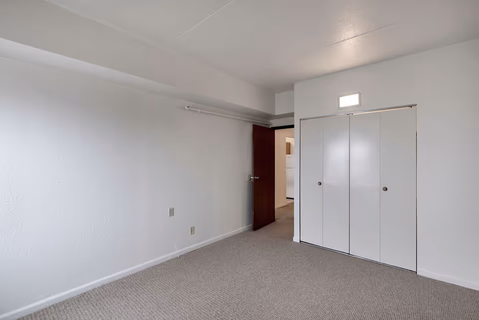 Empty room with beige carpet, white walls, a closed white closet with double doors, and an open dark brown door leading to another room with a visible refrigerator.
