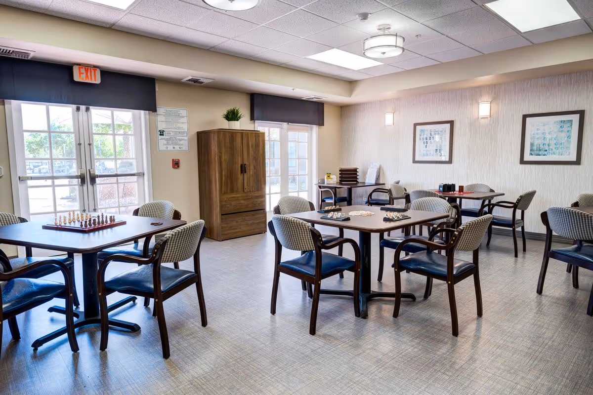 A well-lit common room with several tables and chairs arranged for games and social activities. One table has a chess set, another has a card game setup. The room has large windows and glass doors letting in natural light, framed artwork on the walls, and a wooden cabinet with a plant on top near the exit door.