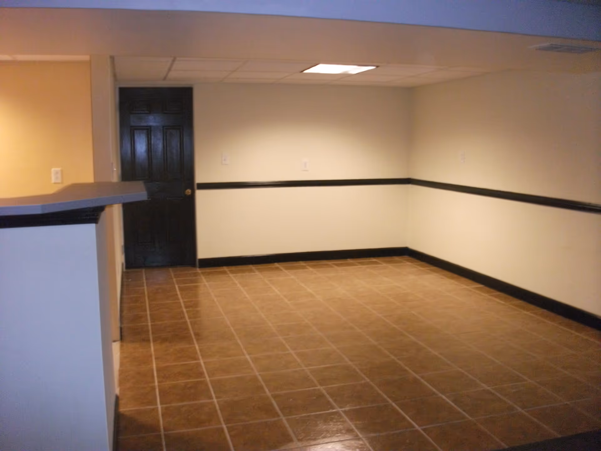 Empty interior room with beige walls, brown tiled floor, black baseboards and chair rail, a black door, and a ceiling light panel.