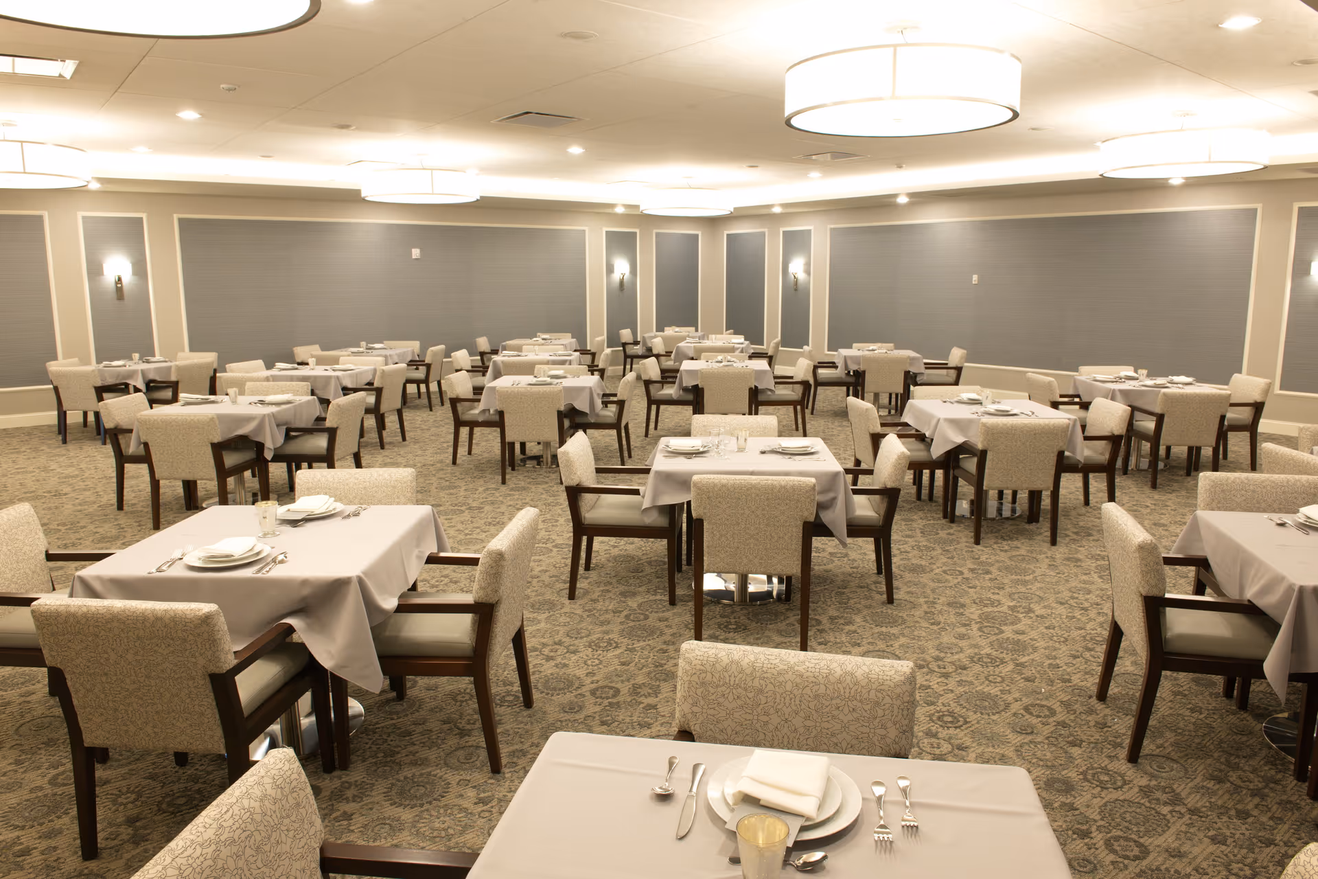 A spacious dining room with multiple tables covered in white tablecloths, each set with plates, napkins, and silverware. The room has beige patterned carpet, upholstered chairs, soft wall lighting, and large ceiling light fixtures.