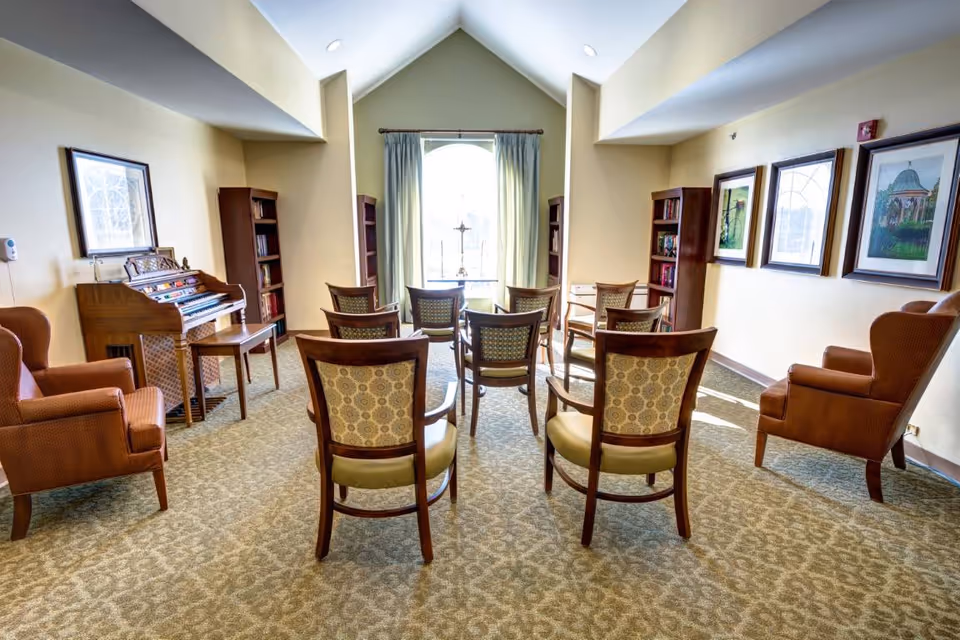 Sunlit communal sitting room with upholstered armchairs and wooden chairs arranged facing a window, with a small organ/piano and framed artwork on the walls.