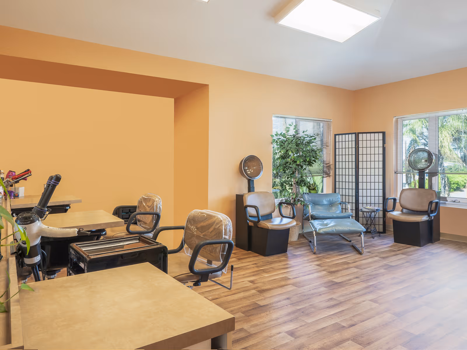 Interior of a salon area with several salon chairs, hair dryers, a blue lounge chair, a folding screen, and large windows with blinds letting in natural light. The walls are painted a warm beige color and the floor has wood-like vinyl flooring.