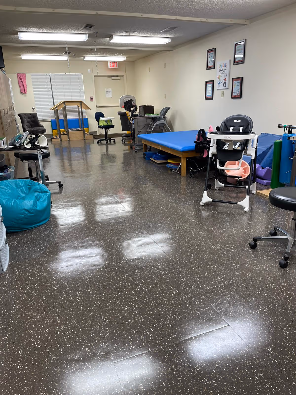A therapy or activity room with various equipment including a blue therapy table, a high chair, chairs on wheels, a bean bag, and a desk with a computer. The room has a speckled floor, fluorescent ceiling lights, framed certificates and a colorful poster on the wall, and an exit door at the far end.
