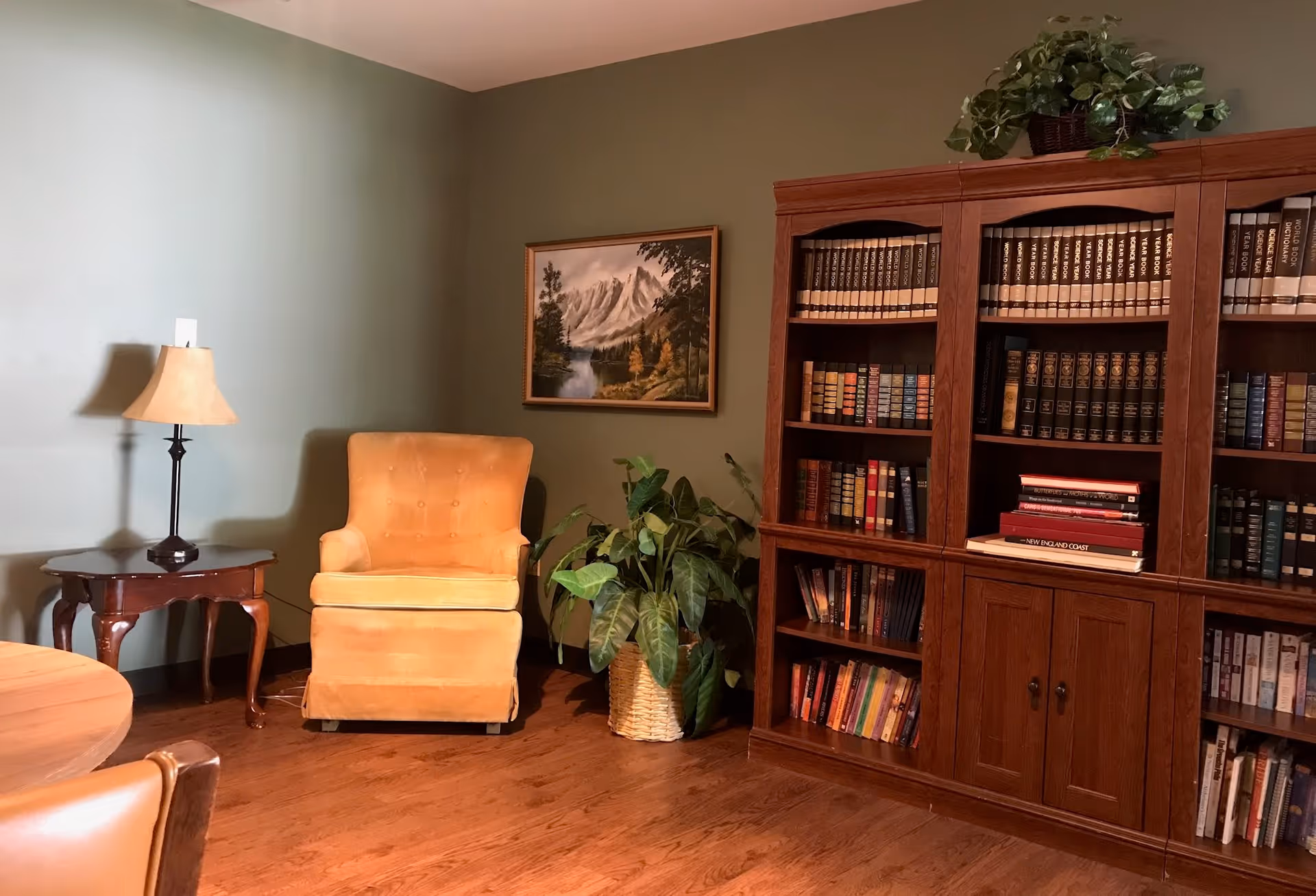 A cozy interior room with a yellow upholstered armchair, a wooden side table with a lamp, a large wooden bookshelf filled with books, a potted plant in a woven basket, and a framed landscape painting of mountains and trees on the wall.