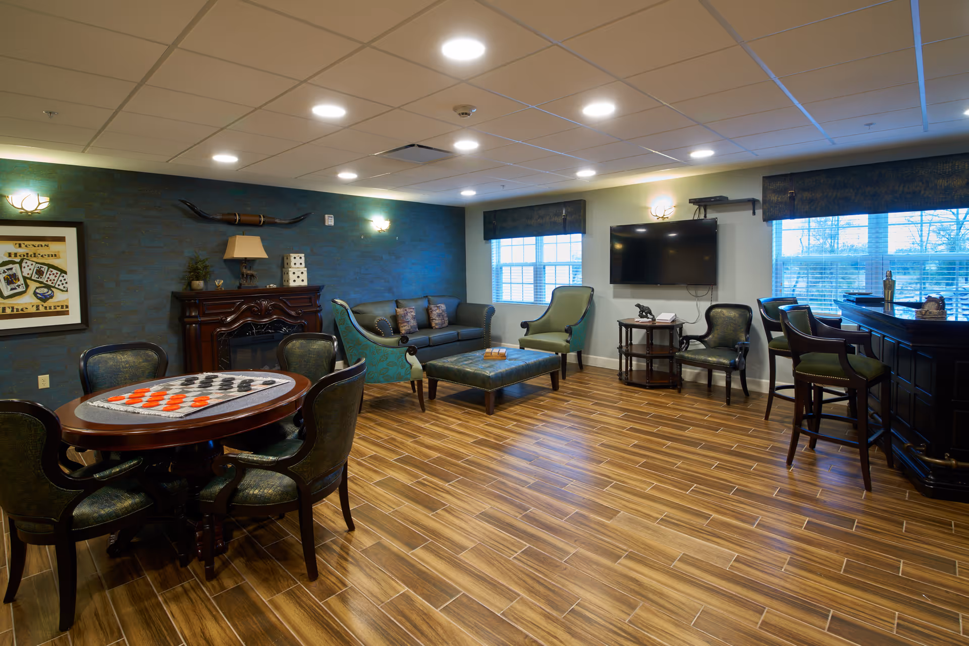 A cozy common area with wooden flooring featuring a round table with a checkers game set up, several upholstered chairs, a sofa, armchairs, a wall-mounted TV, and a bar counter with stools. The room has a blue accent wall with decorative items and windows letting in natural light.