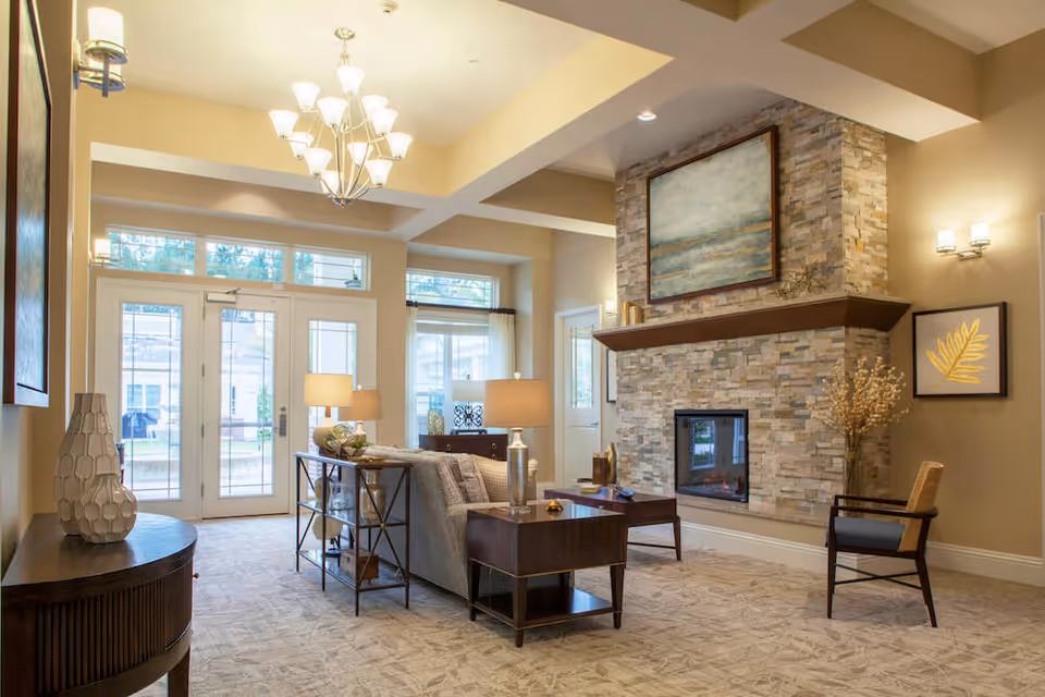 A bright and spacious living room area with a stone fireplace, a large painting above the mantel, a sofa with side tables and lamps, a chair, and large windows and glass doors letting in natural light.