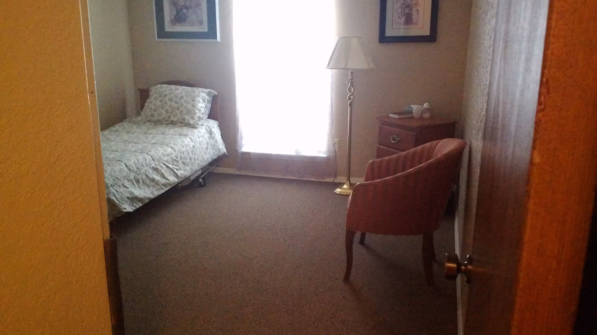 Small bedroom with a single bed against the left wall, a nightstand and floor lamp by a window, and an upholstered chair near the door.