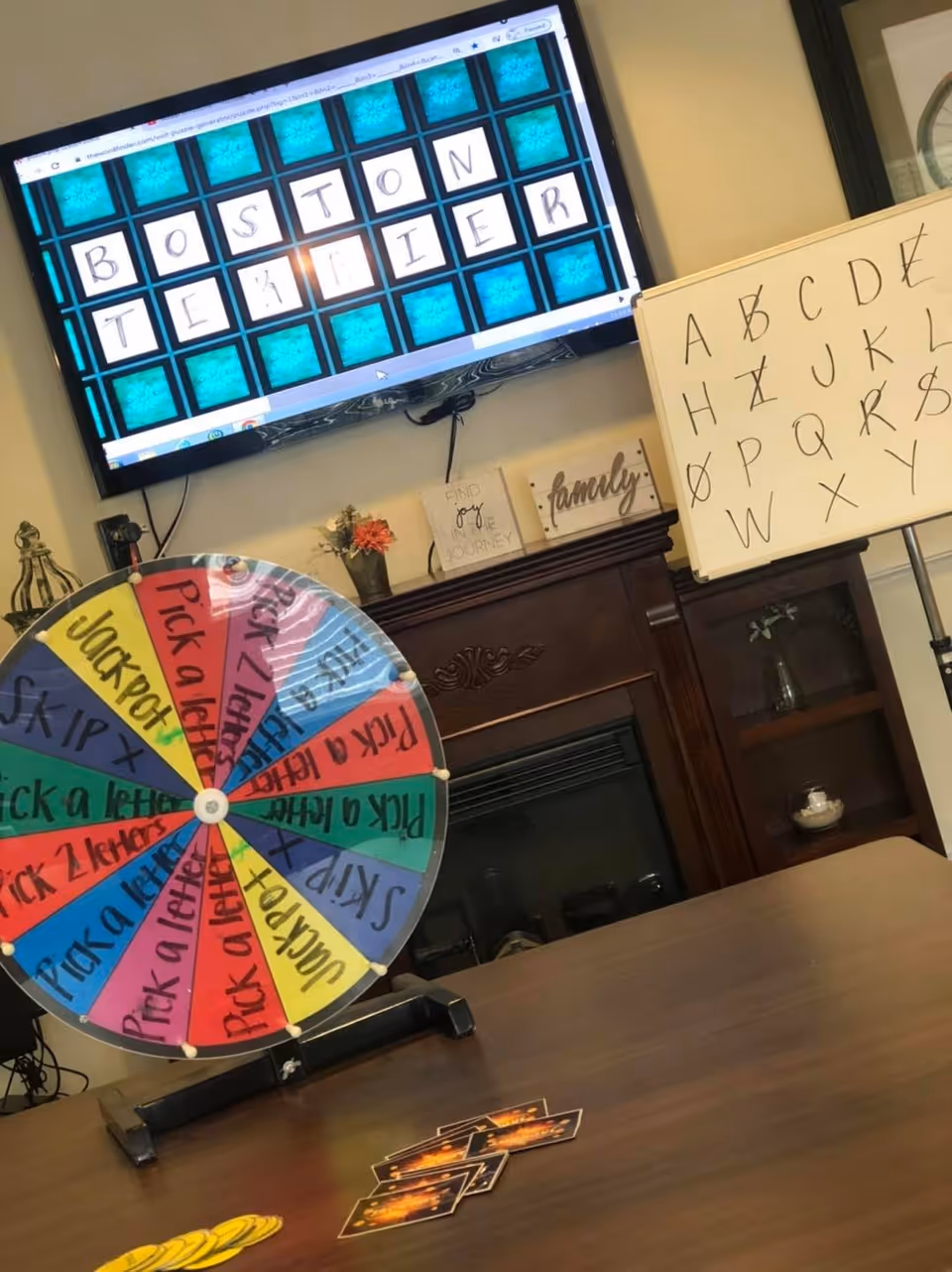 A game setup in a room with a colorful spinning wheel on a table, a whiteboard with crossed-out letters, and a TV screen displaying a word puzzle with the letters spelling 'BOSTON TERRIER'. There are decorative items on a wooden mantelpiece in the background.