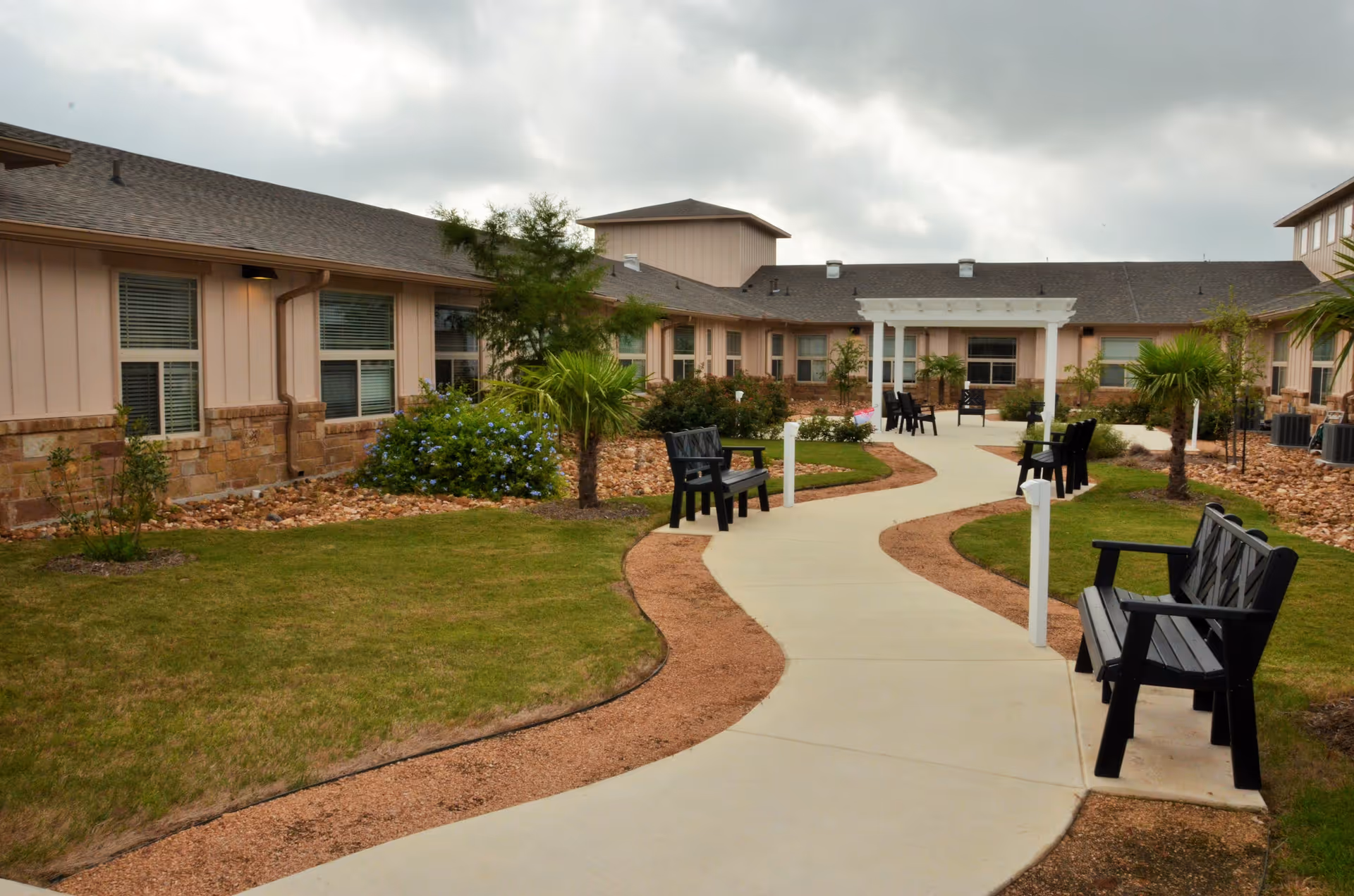 Outdoor courtyard area at The Grandview of Westover Hills featuring a winding concrete pathway, black benches along the path, green grass, landscaped plants, and a building with beige siding and stone accents under a cloudy sky.