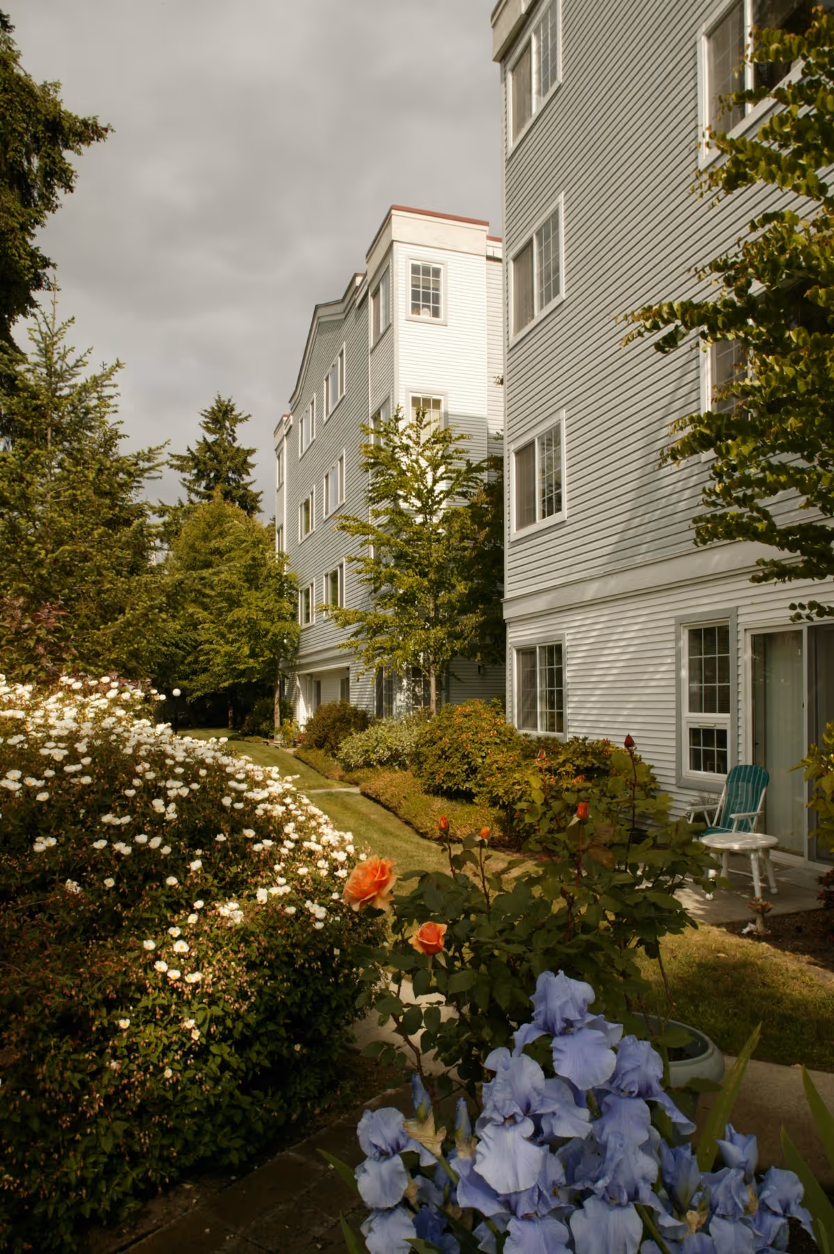 Exterior view of a multi-story light-gray senior living building beside a landscaped garden with blooming flowers and a patio chair.