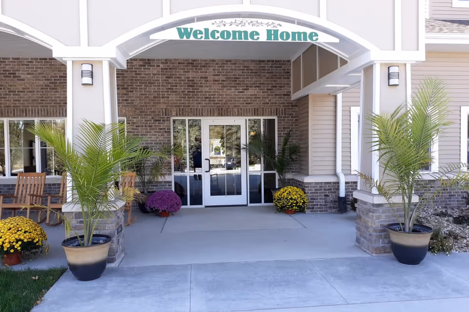 Entrance of a senior living facility with a 'Welcome Home' sign above the doorway. The entrance features glass double doors, brick and siding exterior walls, potted plants, and wooden rocking chairs on the left side.