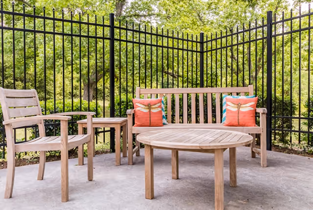 Wooden outdoor seating set with a bench, chairs, round table and colorful pillows by a black metal fence and green trees.