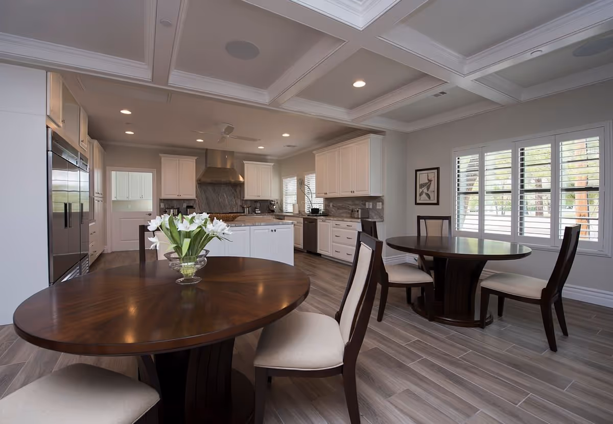 A modern kitchen and dining area with two round wooden tables, each surrounded by chairs with light-colored cushions. The kitchen features white cabinetry, stainless steel appliances, a large range hood, and a central island. The room has large windows with white shutters, recessed lighting, and a coffered ceiling. A vase with white flowers is placed on one of the tables.