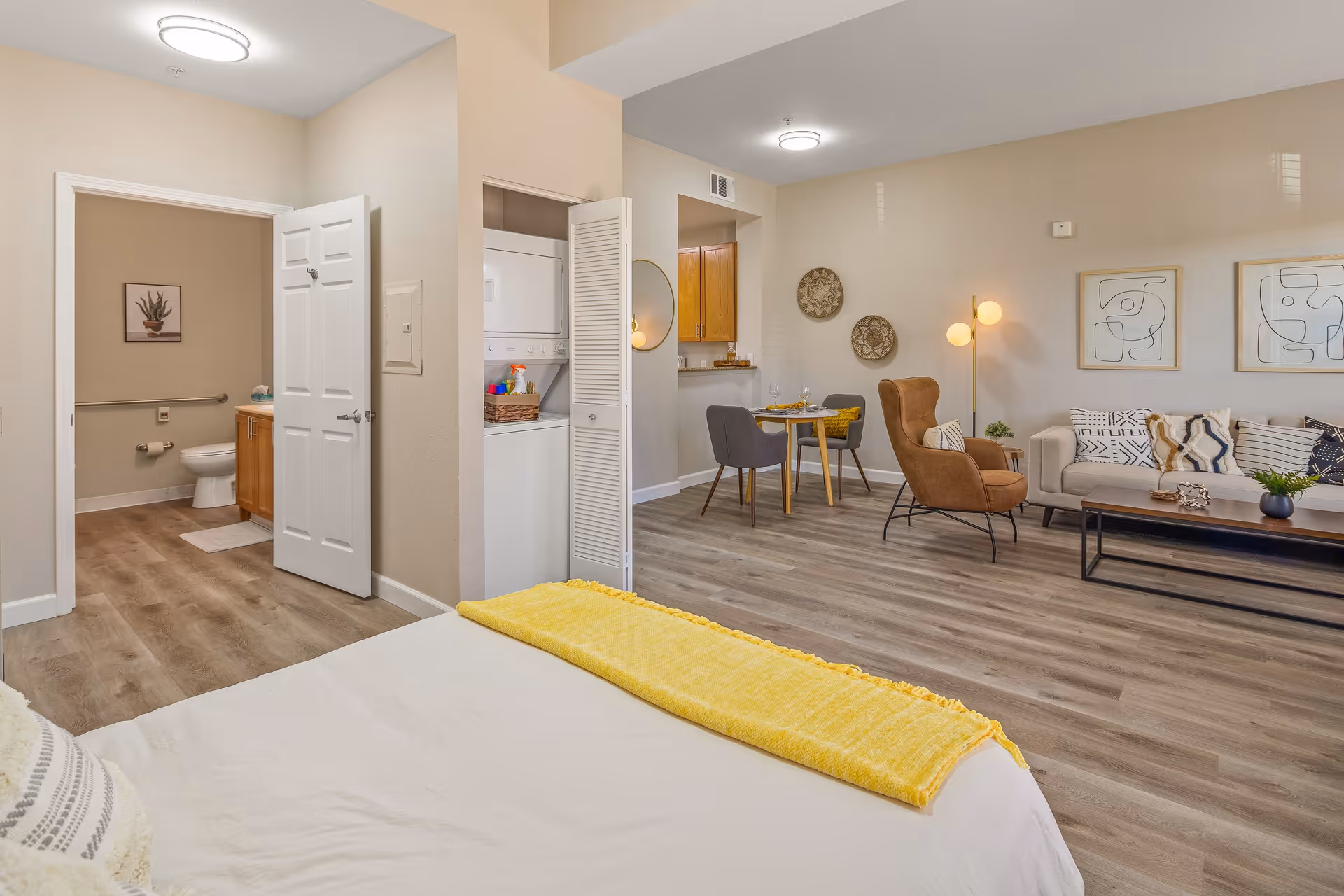 A bright and spacious senior living apartment featuring a bed with a yellow throw blanket in the foreground, an open bathroom with a toilet and vanity visible, a stacked washer and dryer unit in a closet, and a living area with a sofa, armchair, coffee table, dining table with chairs, and modern wall art and decor.