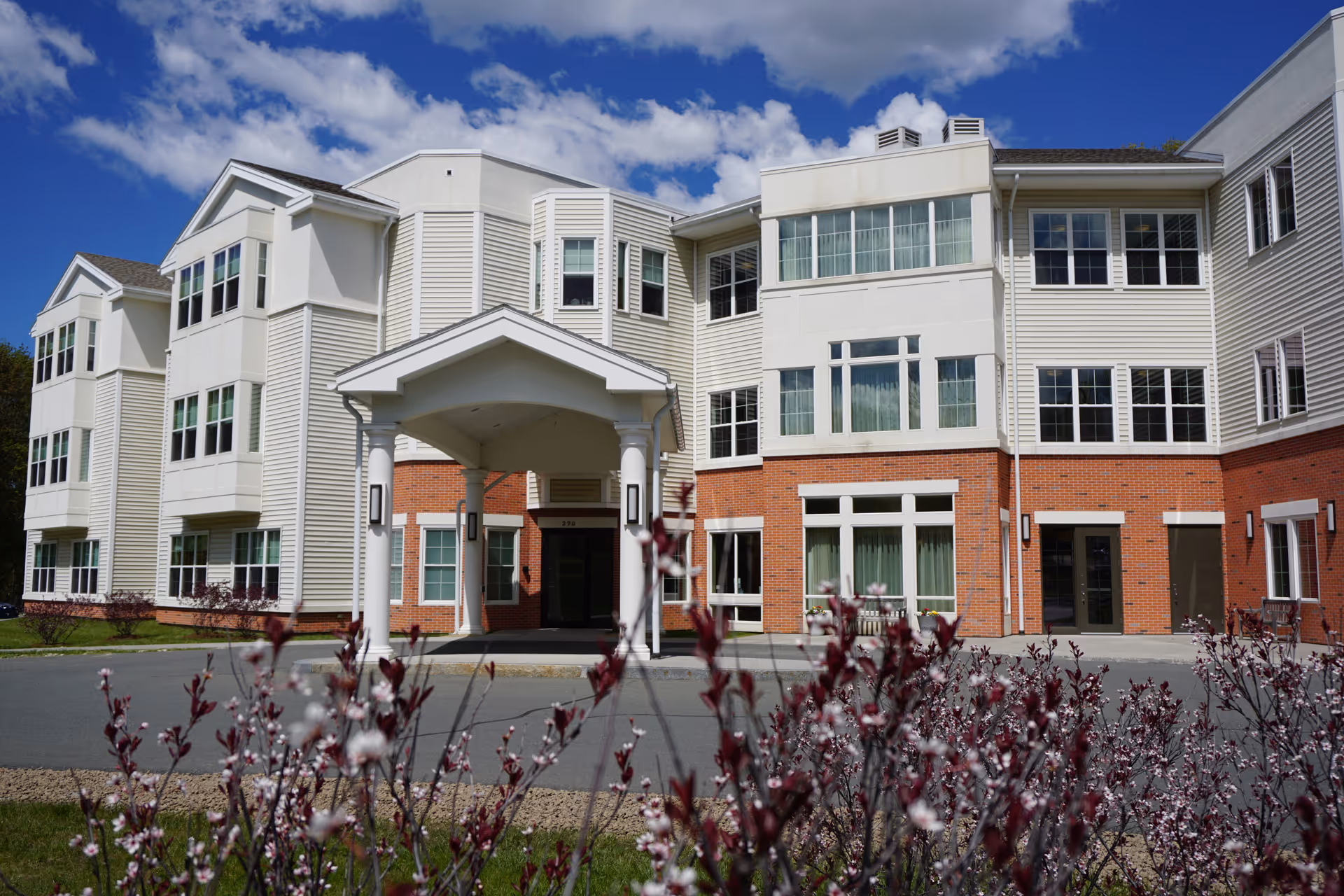 Front entrance of a multi-story senior living building with a covered portico, large windows, and flowering shrubs in the foreground.