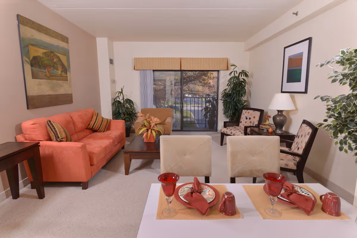 A cozy senior living room with a coral sofa, two beige chairs, a coffee table with a plant, and two patterned armchairs near a side table with a lamp. There is a large window with a view of trees and a street outside. The dining table in the foreground is set with plates, red napkins, red glasses, and mugs.