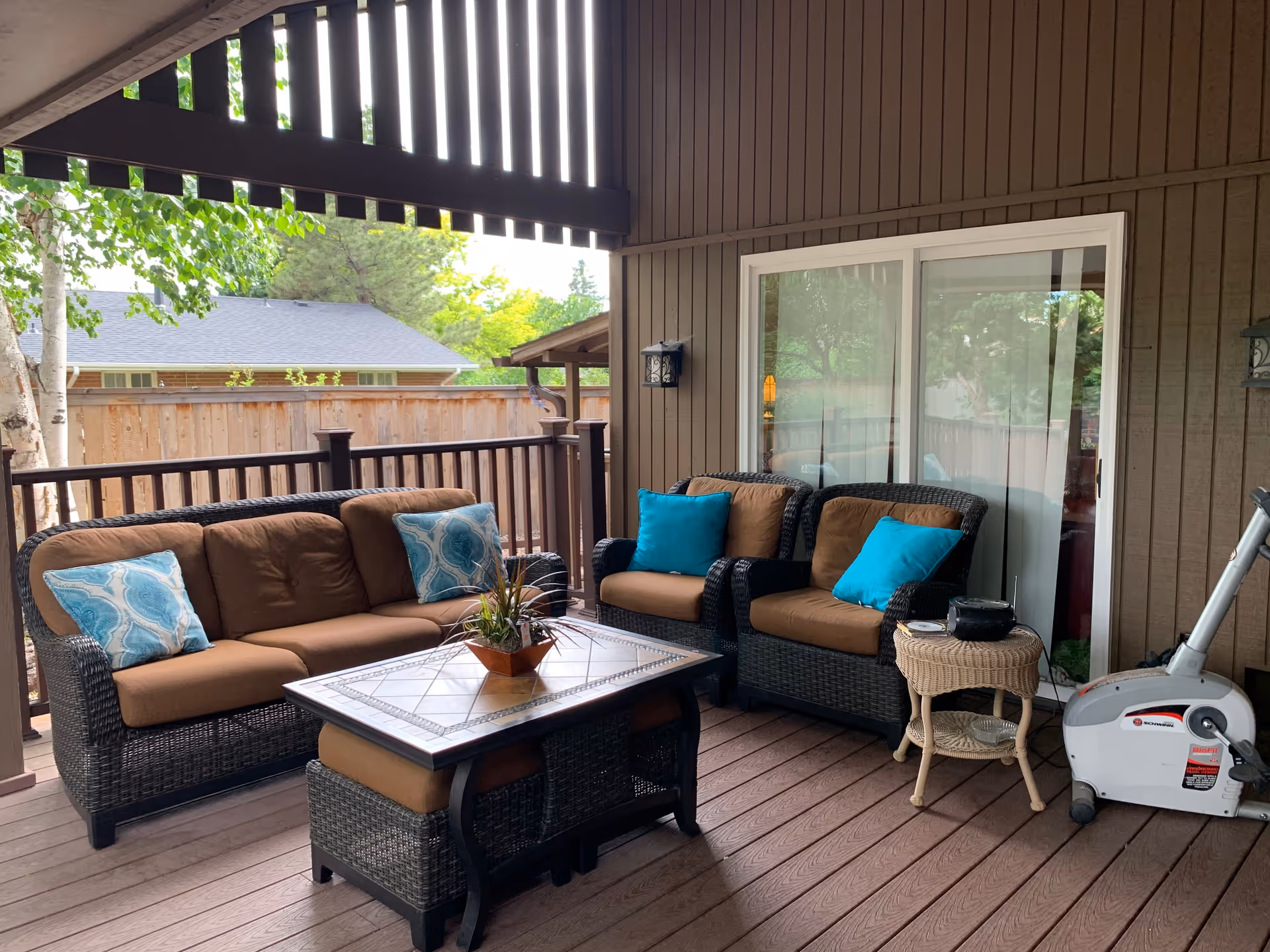 Covered outdoor patio area with brown wicker furniture including a sofa and two armchairs with brown cushions and blue decorative pillows. A rectangular coffee table with a tiled top and a small wicker side table with a black pot on it are also present. There is a stationary exercise bike on the right side. The patio has wooden flooring and railing, with a wooden privacy fence and trees visible in the background.