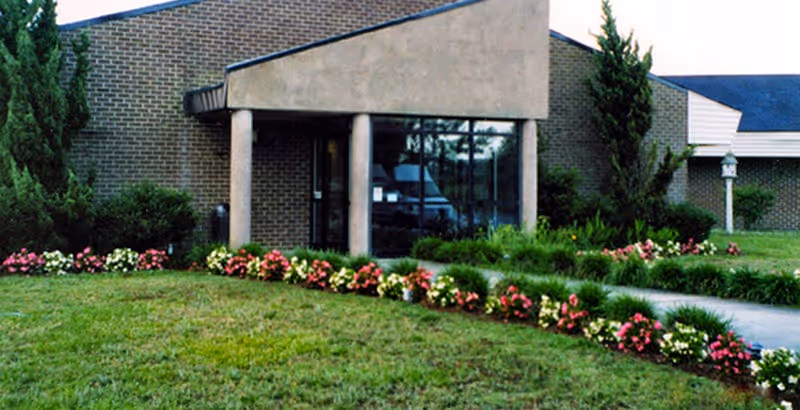 Exterior view of a brick building entrance with two large columns supporting a triangular overhang. The entrance is surrounded by well-maintained landscaping including green grass, bushes, and a row of colorful flowers along the walkway.
