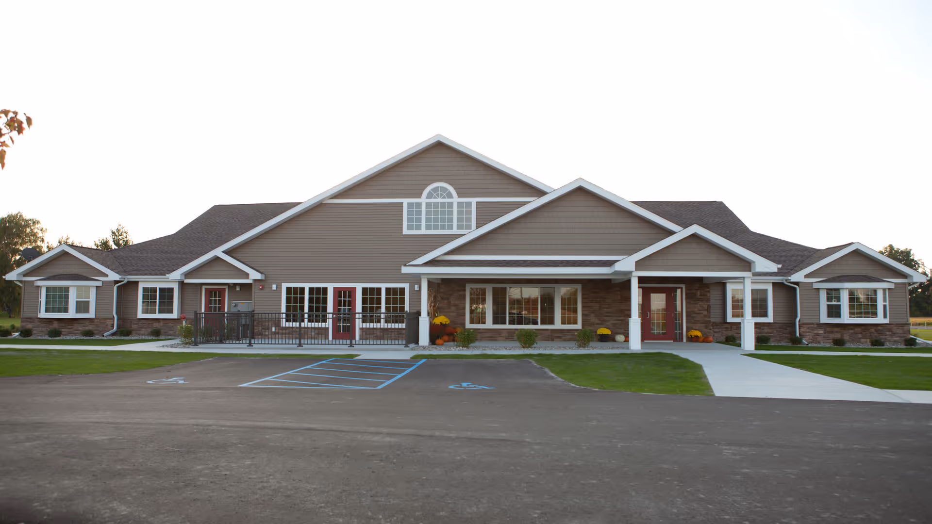 Front exterior of a single-story assisted living building with gabled roofs, large windows, red doors, and a parking area with marked handicapped spaces.