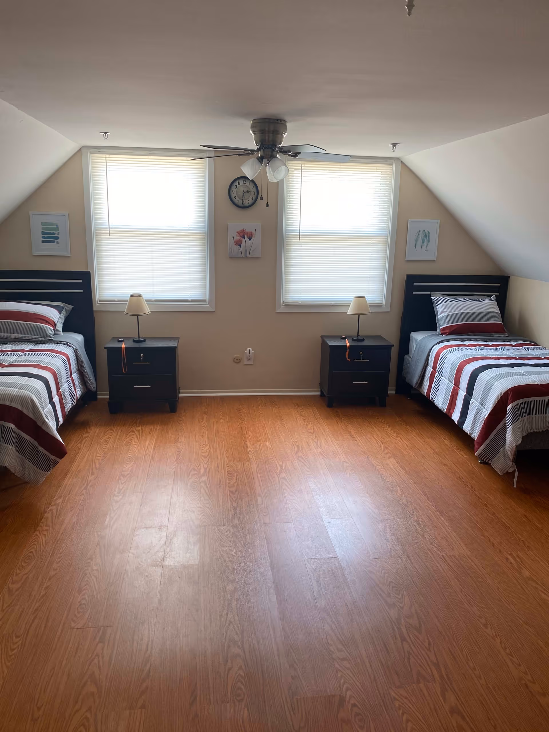 A bedroom with two single beds placed against opposite walls, each with striped bedding in shades of red, gray, and white. Between the beds are two black nightstands, each with a small lamp. Two windows with blinds are centered on the far wall, with a clock and framed artwork hanging between and beside them. The room has wooden flooring and a ceiling fan with lights.