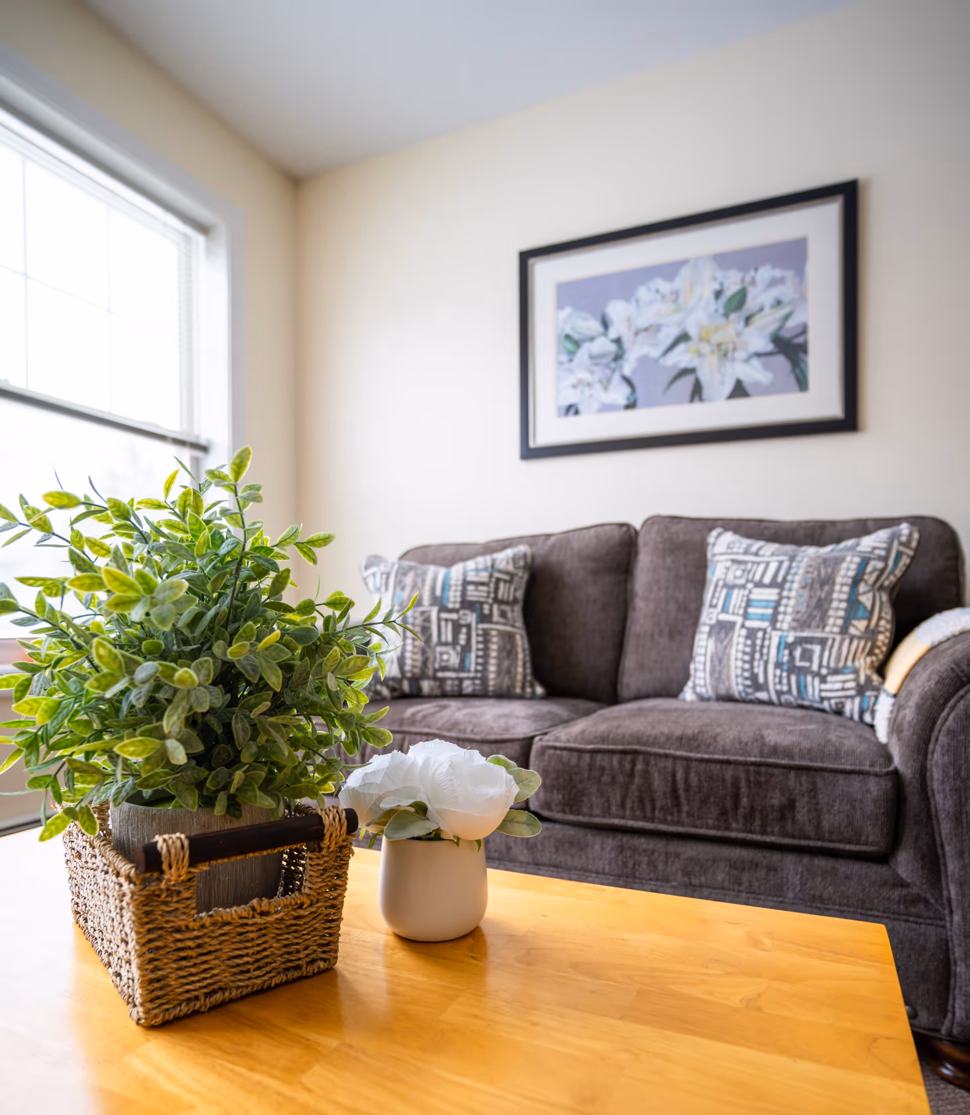 A cozy living room with a dark gray sofa adorned with patterned pillows. In front of the sofa is a wooden coffee table with a small potted plant in a woven basket and a white flower in a small white vase. A framed floral artwork hangs on the wall above the sofa, and a large window allows natural light to brighten the space.