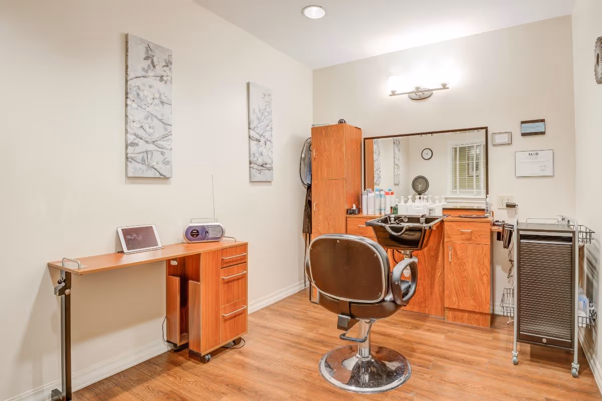 A small hair salon room with a black salon chair in front of a wooden cabinet and sink. The cabinet has various hair care products on top. There is a large mirror above the cabinet, two floral paintings on the wall, a small wooden desk with a tablet and a radio, and a rolling cart with supplies on the right side. The room has wooden flooring and white walls.