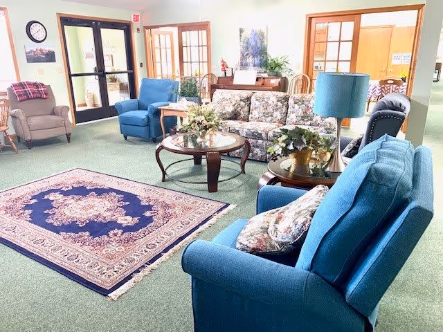 Bright communal lounge with upholstered armchairs and a floral sofa arranged around coffee tables and a patterned rug.