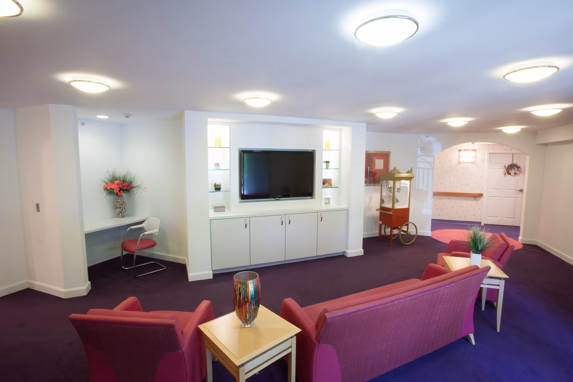 A cozy living room area in a senior living facility with purple carpet and pink upholstered chairs and sofas arranged around light wood side tables. A flat-screen TV is mounted on a white built-in cabinet with shelves displaying small decorative items. There is a small desk with a chair and a vase of flowers in the corner. A vintage popcorn machine is positioned near an arched doorway leading to a hallway with a white door and a wreath.