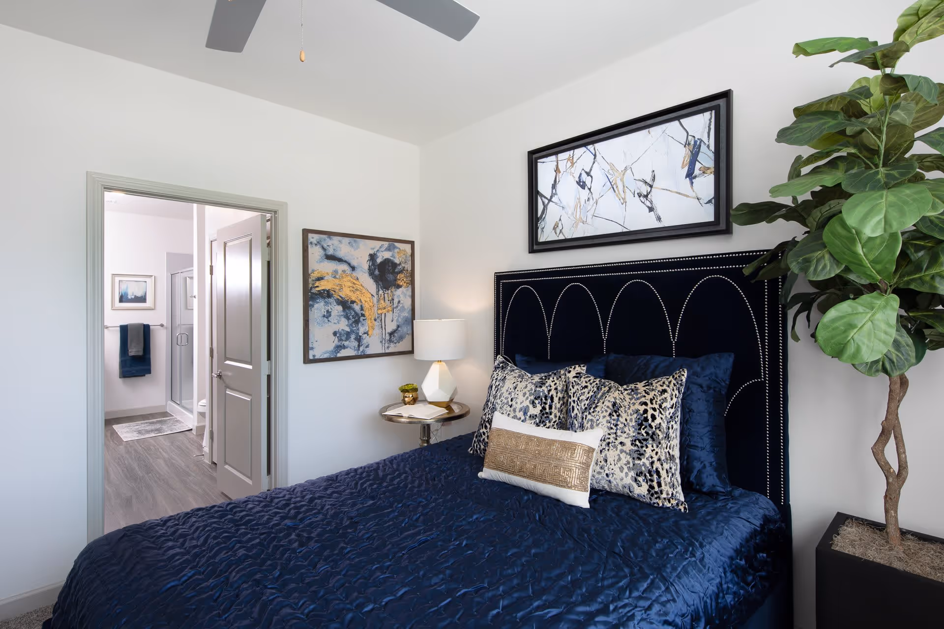 A bedroom with a navy blue bedspread and decorative pillows on a bed with a dark upholstered headboard. There is a small round side table with a lamp and a small plant next to the bed. Two abstract paintings hang on the white walls, and a large green potted plant is in the corner. An open door reveals a bathroom with a glass shower and towels hanging on the wall.