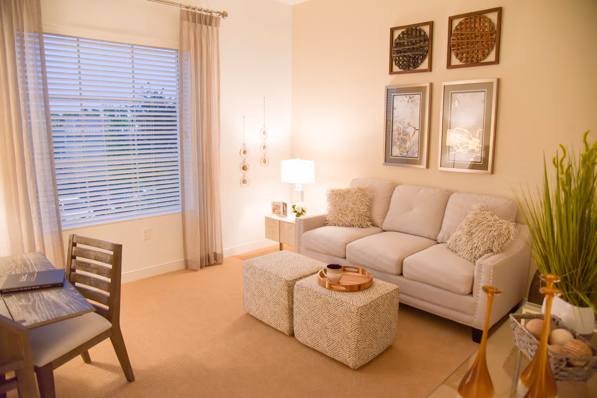 A cozy living room with a beige sofa adorned with two fluffy pillows, two patterned ottomans with a wooden tray on top, a small side table with a lamp and decorative items, a wooden chair and desk, beige carpet, large window with blinds and curtains, and wall art including two framed pictures and two round woven pieces.