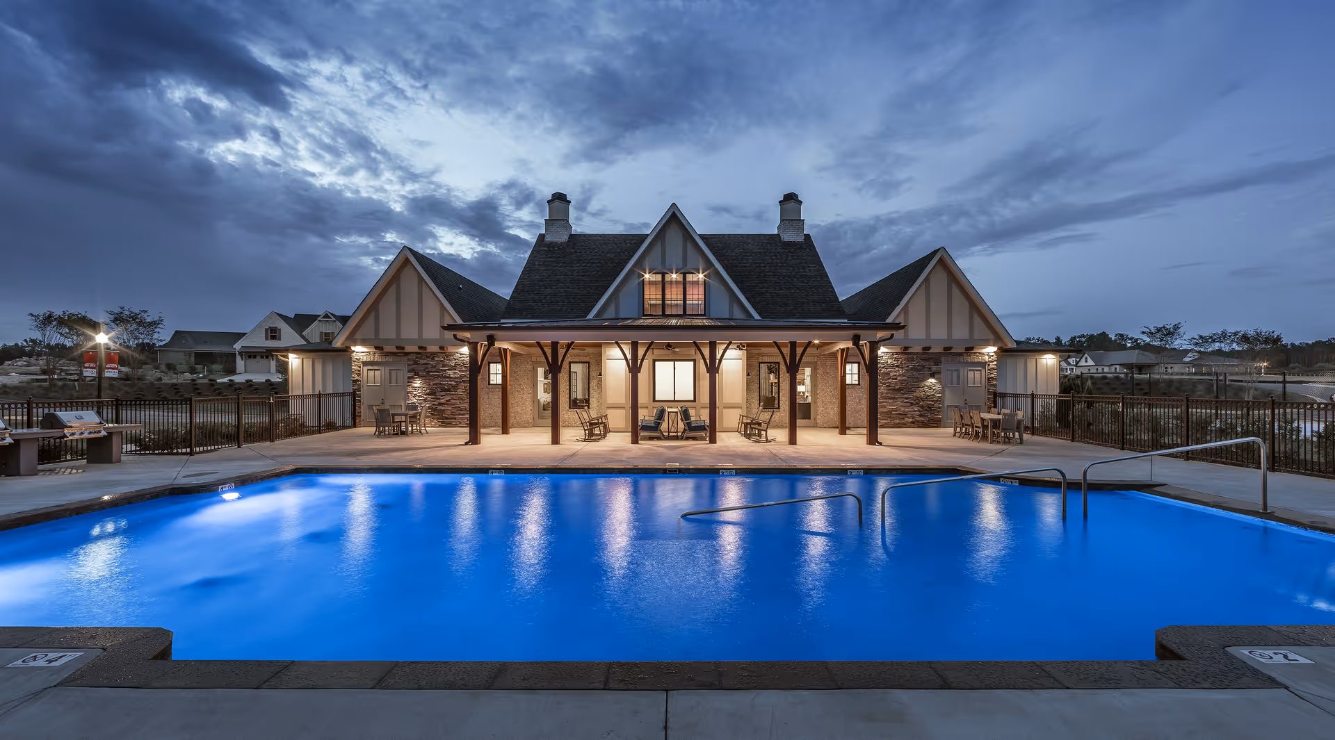 Evening view of a well-lit swimming pool in front of a clubhouse building with a steep roof and wooden pillars, surrounded by a fenced area and outdoor seating.
